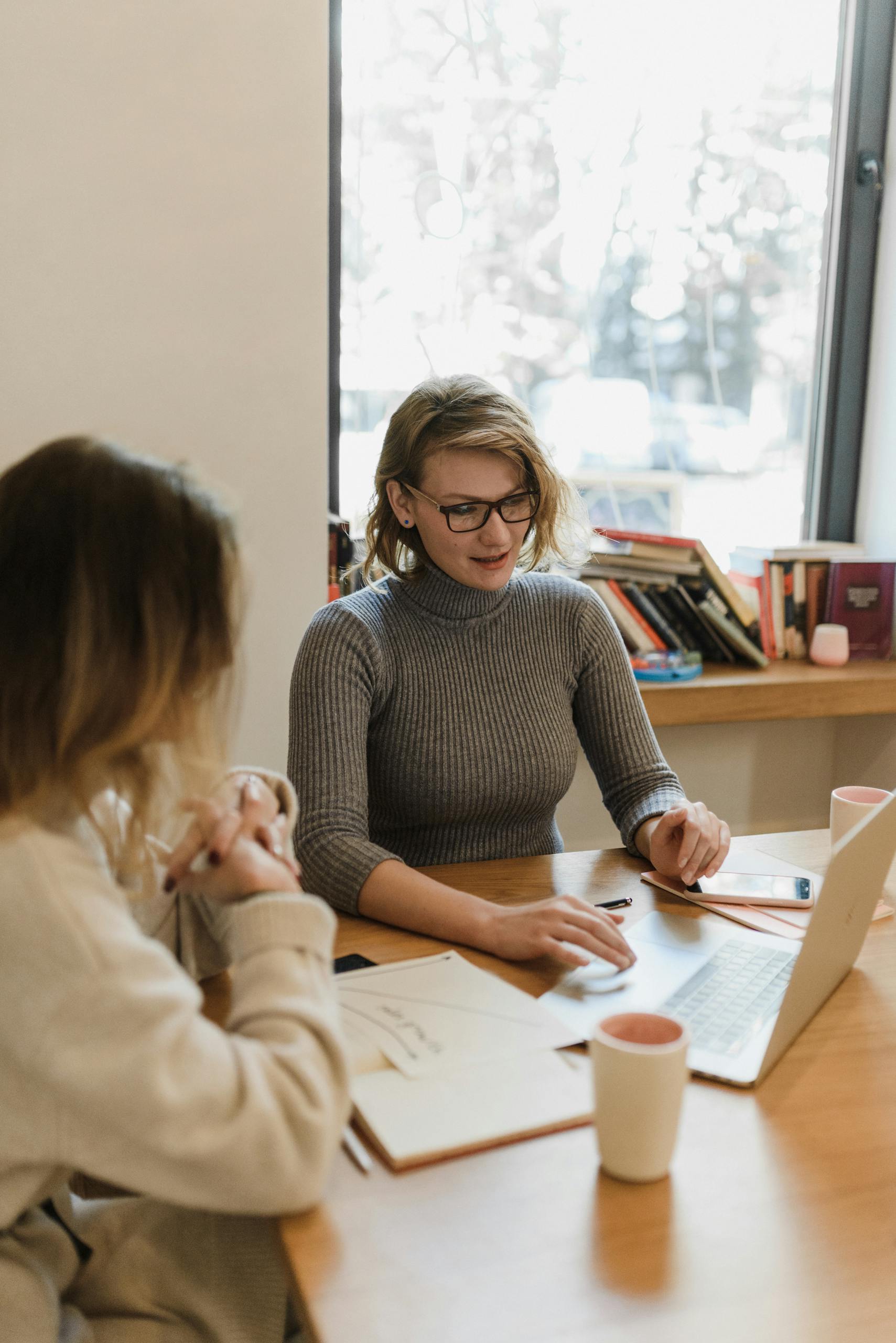 Two women working together on a laptop in a well-lit office setting, discussing ideas.